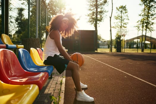 Attractive confident young sportswoman with basketball ball in hands sitting taking rest during training class at outdoor court