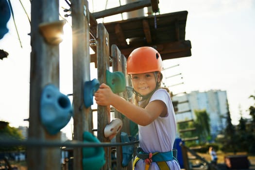 City outdoor rope park for happy childhood and summer leisure activity. Portrait of satisfied girl child climbing with protective equipment feeling fun and courage