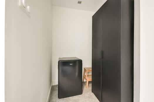Interior view of a small room featuring a black fridge, a wardrobe, and a wooden chair. The minimalist design adds a modern touch to the space.