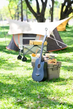 An acoustic guitar leaning on picnic basket at a forest campsite.