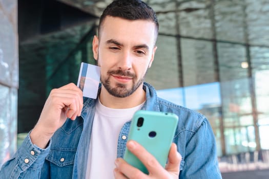 Happy young man using his smartphone while holding a credit card. He smiles with excitement, standing outside a glass building, enjoying an online shopping or payment experience.