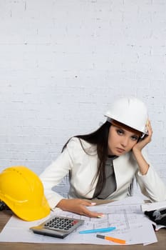 A stressed engineer reviews construction plans at a cluttered desk, wearing a hard hat with tools and documents