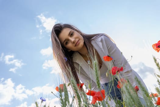 Smiling brunette girl is looking down at the camera in a field of red poppies. The blue sky is in the  background.. Horizontally.