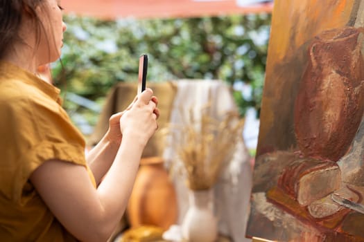 Confident painter presenting her new masterpiece on online branding video, filming a vlog on smartphone to promote the artistic products used for the artwork on canvas. Art and craft content creation.