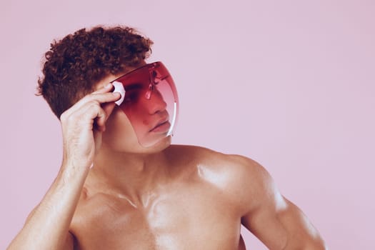 Stylish young man with curly hair and no shirt, playfully holding pink sunglasses. The soft pink background enhances the fashion-forward vibe and individual expression.