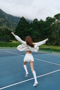 A young athletic female in white sportswear exuberantly running on a tennis court, highlighted by a vibrant green mountain landscape, conveying themes of energy, sports, and nature