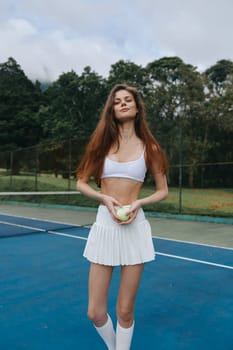 Energetic young woman in a white sports outfit, confidently holding a tennis ball on a blue court, surrounded by green trees, symbolizing fitness and athleticism