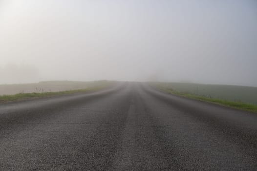 An empty asphalt road fades into dense morning fog, with fields and sparse trees barely visible in the misty distance.