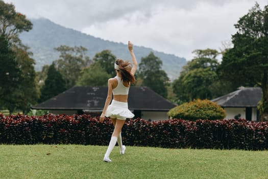 A cheerful young woman in a white outfit twirls in a lush green park, surrounded by nature and mountains, radiating joy and freedom