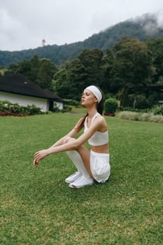 Asian woman in a sporty white outfit and headband squats on grass, embodying fitness and tranquility in a green outdoor setting, perfect for wellness themes