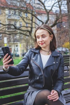 Young woman sitting on a park bench taking a selfie using her phone. High quality photo