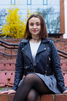 Cheerful student girl in a black leather jacket and short skirt sitting on the street with her legs crossed. High quality photo