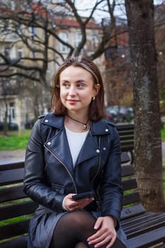 A young woman with a phone in her hands sits on a park bench and waits for a meeting. High quality photo