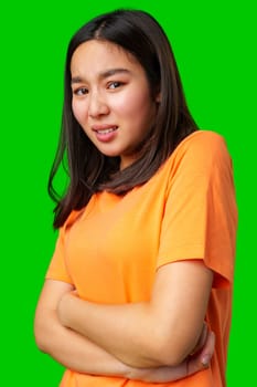 A young woman stands with her arms crossed, wearing an orange shirt. Her expression conveys confusion or uncertainty, against a striking green background.