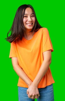 A young woman stands cheerfully with her hands clasped in front of her, wearing a casual orange shirt and denim shorts against a vivid green background. Her joyful expression radiates positivity.