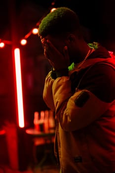 A figure stands under red lights, covering their face in a moment of deep thought or emotion. The urban backdrop adds to the intensity of the night atmosphere.