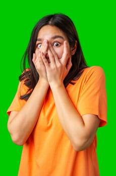 A woman in an orange shirt shows a shocked expression, covering her mouth with both hands while staring wide-eyed. The bright green background adds to the playful atmosphere.