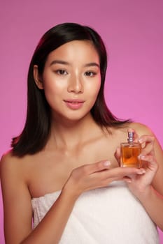 A young woman poses against a soft pink backdrop, elegantly holding a bottle of perfume. She appears fresh and natural, showcasing her radiant skin.