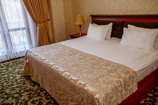 Hotel room with golden yellow blanket on dark red bed and green carpet on the floor. Room in golden beige tones with white bed linen. on the bedside table table lamp