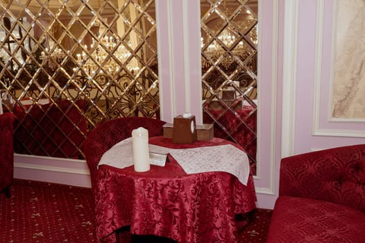 A round table in a restaurant, covered with a red tablecloth, next to it a wall with mirrors. On the table are napkins, a candle and a box with paper napkins.