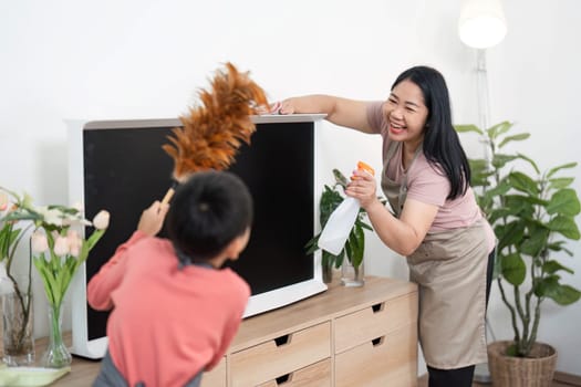 A mother and her son share a joyful moment while cleaning a TV stand, demonstrating teamwork and teaching valuable life skills in a cozy home environment.