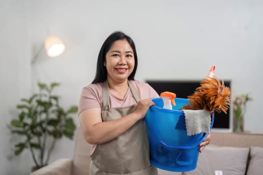 A happy woman stands in her living room with a bucket of cleaning supplies, ready to maintain a spotless home environment.