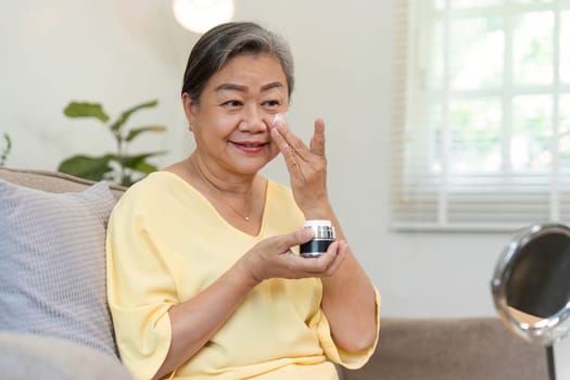 A joyful senior woman takes a moment for herself, applying moisturizer while enjoying her skincare routine in a cozy living space, highlighting the importance of self-care.