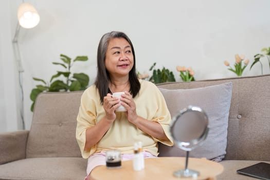 A confident older woman embraces her beauty routine, holding a cup of tea and reflecting on self-care in a serene home environment, showcasing age diversity and wellness.