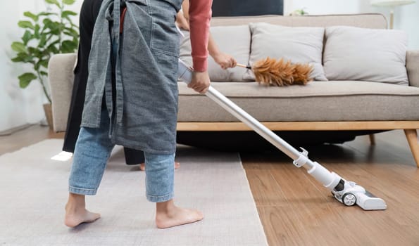 A young boy enthusiastically vacuums the floor, while his mother helps by dusting the furniture, creating a fun and collaborative cleaning environment.