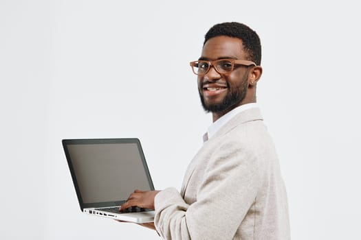 Happy man with glasses holding a laptop, dressed in a beige blazer and white shirt, smiling confidently, facing camera, studio setting with plain white background, representing technology, business, confidence, and professionalism, concept: modern work culture