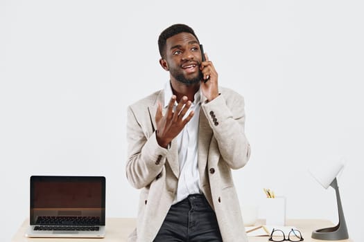 Cheerful young African-American man in casual beige cardigan smiling and talking on mobile phone in modern office environment with laptop, glasses, and stationery on clean white desk, expressing positivity. Concept: professional communication.