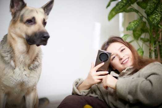 A young woman sits on the floor, smiling while using her smartphone next to her German Shepherd dog in a warm, inviting room.