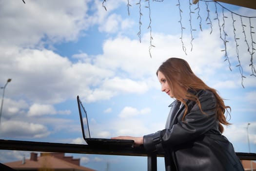 A young woman is engaged in work on her laptop at an outdoor location with a clear blue sky.