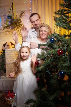 A family gathers in a warm room, smiling and waving near a festive Christmas tree adorned with colorful decorations and lights.