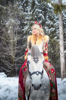 A stunning Russian girl dressed in vibrant national attire enjoys a photoshoot with a horse amidst snowy scenery.