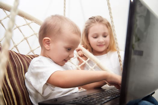 Two children engage with a laptop in a hammock, showcasing curiosity and connection in a relaxed indoor setting.