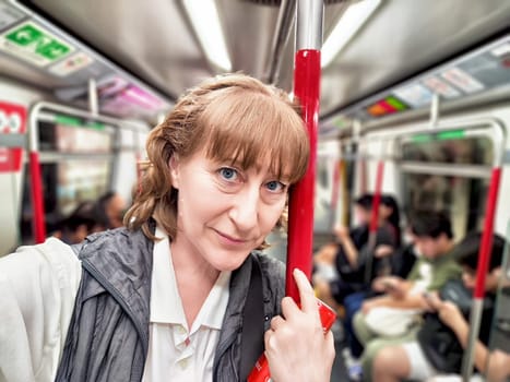 A middle-aged blonde woman enjoys a subway ride, capturing a selfie while surrounded by fellow commuters, immersed in urban exploration.