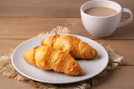 Two golden croissants rest on a white plate beside a steaming cup of coffee. The warm wooden table adds a rustic touch to this inviting breakfast arrangement.