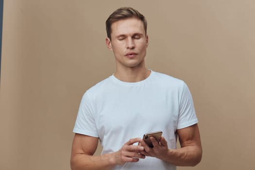 Casual young man with short brown hair and fair skin wearing a white T-shirt, holding a smartphone and focused on screen, standing in front of a beige wall. Concept: modern technology, communication, lifestyle, relaxed mood, indoor portrait, youth.