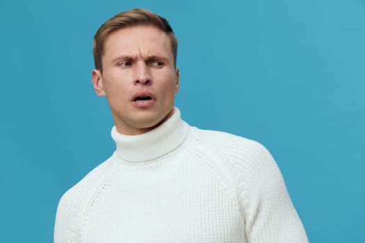 Close-up of a man with questioning and confused look, wearing white turtleneck sweater against a plain blue background. Studio shot emphasizing facial expression and emotion. Concept: confusion, doubt