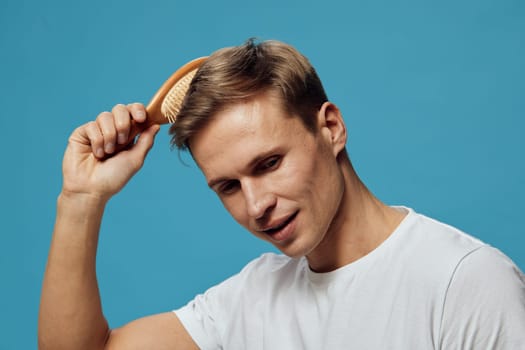 Close-up of a young man with short brown hair using a wooden comb to tidy his hairstyle against a bright blue backdrop. He wears a simple white t-shirt, capturing a personal grooming moment, emphasizing cleanliness and self-care. Concept: grooming routine.