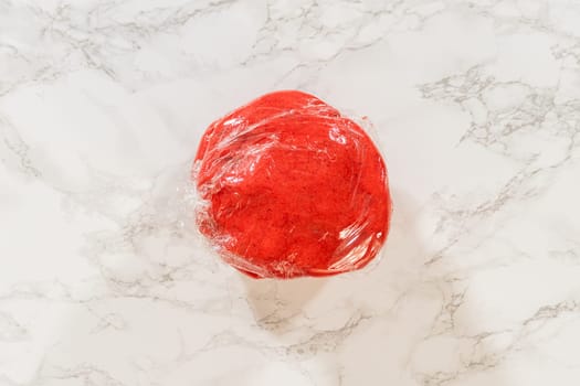 Close-up of a ball of Classic Red Gingerbread Dough wrapped in plastic wrap, highlighting its vibrant red color and readiness for chilling.