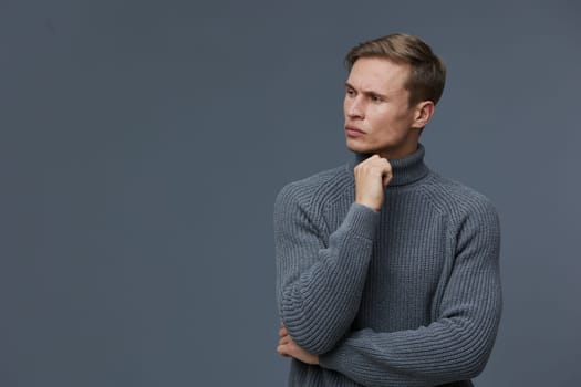 Confident man wearing a cozy gray sweater appears deep in thought against a plain gray backdrop. The studio shot emphasizes his contemplative mood and casual style, capturing a moment of introspection and focus, ideal for lifestyle and emotion themes.