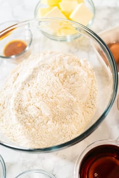 Side view of various baking ingredients arranged around a large glass bowl filled with flour on a marble surface. Ingredients include butter, eggs, brown sugar, spices, and maple syrup, showcasing the setup for Amber Gingerbread Cookie Dough.