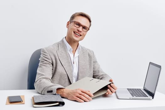 smiling man working with laptop at desk dressed in gray blazer and glasses using digital technology for freelance remote job as digital nomad concept with smartphone and notebook nearby
