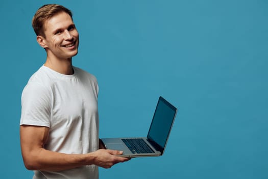 smiling young caucasian man hold laptop in hands look camera wear casual white t-shirt isolated on blue background remote work digital nomad concept technology and freelancing lifestyle