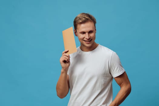 Happy young man with fair hair and casual white t-shirt, smiling while holding a blank cardboard placard to his head against a vibrant blue backdrop. Studio portrait with a positive, cheerful vibe. Concept: personal advertising, promotion, communication, positivity, youthful energy