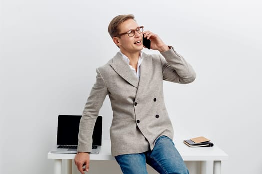 Smiling young caucasian man working and talking on phone wearing gray jacket and jeans sitting on white table with open laptop and notebook beside him. Digital nomad concept of remote work communication and connectivity.