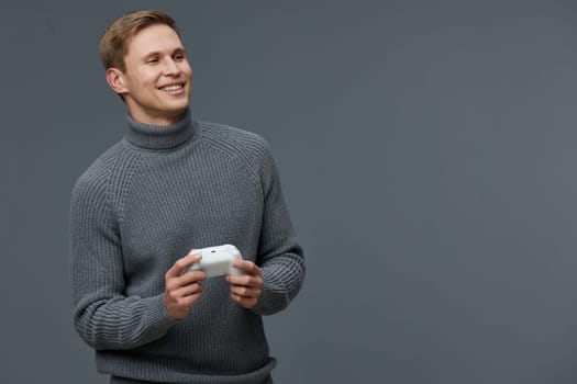 Cheerful young man with light brown hair and a bright smile, dressed in a warm gray turtleneck, enjoying gaming with a controller in his hands. The simple gray background highlights his joyful expression and relaxed atmosphere. Concept: leisure and entertainment.