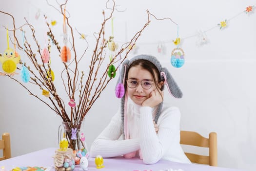 Portrait of one beautiful Caucasian girl in glasses, white turtleneck and bunny headband, who is sitting at the table with homemade Easter bouquet of pussy willow branches in a jug on a spring day, side view close-up. Concept of happy childhood, happy Easter, children leisure, creative work.